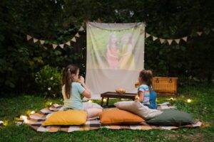 Two girls snacking and watching a projector in the backyard on pillows and a blanket at a party.